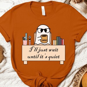 May include: Orange t-shirt with a ghost wearing glasses and holding a coffee mug. The ghost is sitting at a desk with books and pencils. The text on the shirt reads "I'll just wait until it's quiet."