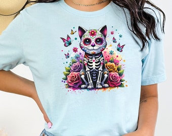 May include: A light blue t-shirt with a colorful graphic of a skeleton cat with a sugar skull face, surrounded by flowers and butterflies.