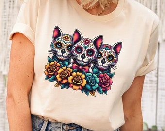 May include: A cream colored t-shirt with a graphic of three sugar skull cats with floral accents. The cats are wearing bow ties and have colorful flowers around them.