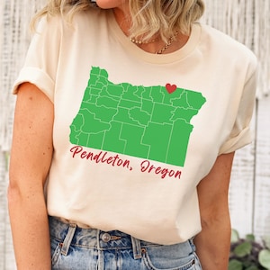 Puede incluir: Una camiseta beige con un contorno verde del estado de Oreg&oacute;n y un coraz&oacute;n rojo que marca la ubicaci&oacute;n de Pendleton, Oreg&oacute;n. El texto "Pendleton, Oreg&oacute;n" est&aacute; escrito en rojo debajo del mapa.