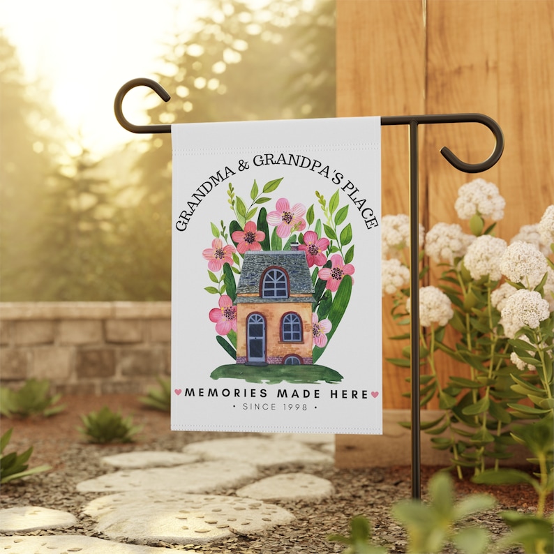 May include: A white garden flag with a watercolor illustration of a house with pink flowers and green leaves. The text on the flag reads "Grandma & Grandpa's Place. Memories Made Here. Since 1998."