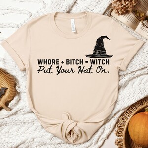 May include: A beige short-sleeve t-shirt featuring a black graphic of a witch hat and the text "WHORE + BITCH = WITCH Put Your Hat On." The shirt has a crew neckline and a standard fit, with the design printed in a bold, dark font across the chest area.