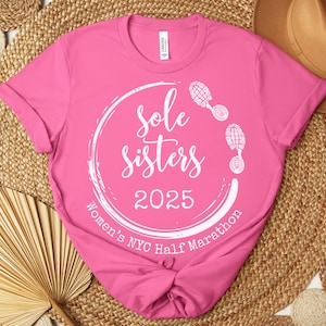 May include: Pink t-shirt with a white circle design that says "Sole Sisters 2025 Women's NYC Half Marathon" with two white footprints on the top and right side of the circle.