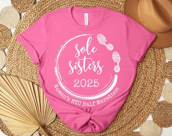 May include: Pink t-shirt with a white circle design that says "Sole Sisters 2025 Women's NYC Half Marathon" with two white footprints on the top and right side of the circle.