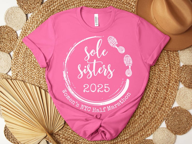 May include: Pink t-shirt with a white circle design that says "Sole Sisters 2025 Women's NYC Half Marathon" with two white footprints on the top and right side of the circle.