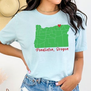 Puede incluir: Una camiseta azul claro con un contorno verde del estado de Oreg&oacute;n y un coraz&oacute;n rojo sobre Pendleton. El texto "Pendleton, Oregon" est&aacute; escrito en rojo cursivo debajo del mapa.