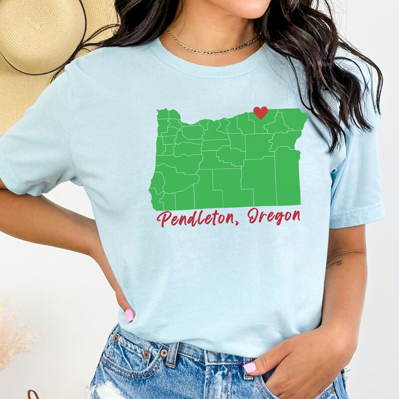 Puede incluir: Una camiseta azul claro con un contorno verde del estado de Oreg&oacute;n y un coraz&oacute;n rojo sobre Pendleton. El texto "Pendleton, Oregon" est&aacute; escrito en rojo cursivo debajo del mapa.