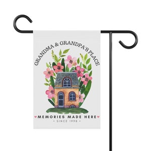 May include: A white garden flag with a house illustration surrounded by pink flowers and green leaves. The text reads "Grandma & Grandpa's Place. Memories Made Here. Since 1998."
