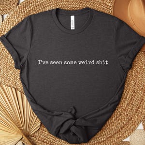 May include: Dark gray t-shirt with the text "I've seen some weird shit" in white. The shirt is displayed on a woven background with a brown hat and decorative fan.
