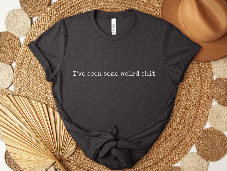 May include: Dark gray t-shirt with the text "I've seen some weird shit" in white. The shirt is displayed on a woven background with a brown hat and decorative fan.