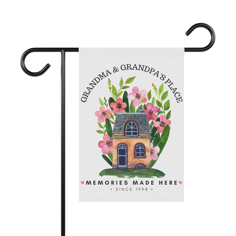 May include: A white garden flag with a watercolor illustration of a house with pink flowers and green leaves. The text on the flag reads "Grandma & Grandpa's Place Memories Made Here - Since 1998".
