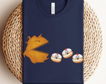 May include: Navy blue t-shirt featuring a cartoon monster chasing three donuts with surprised faces. The monster is orange with sharp teeth. The donuts have white frosting and a red cherry on top. The shirt is folded on a woven surface.