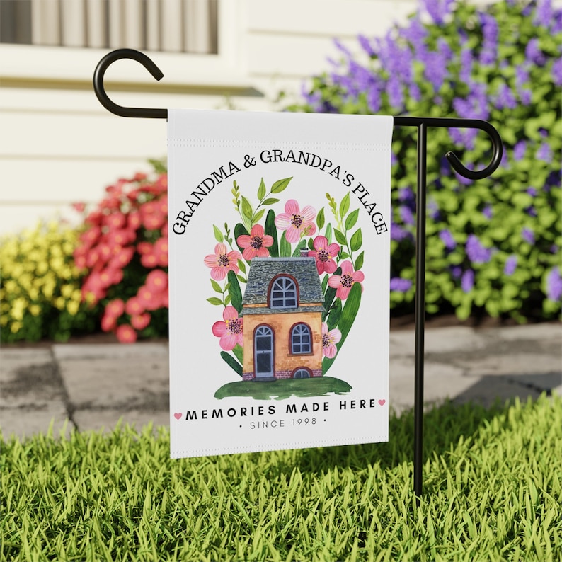 May include: A white garden flag with a watercolor illustration of a house with pink flowers and green leaves. The text on the flag reads "Grandma & Grandpa's Place" and "Memories Made Here - Since 1998 - ".