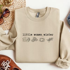 May include: Beige sweatshirt with the text "little women winter" and illustrations of books, oranges, a cup of coffee, and a sprig of greenery. The sweatshirt is paired with jeans, sunglasses, and brown shoes.