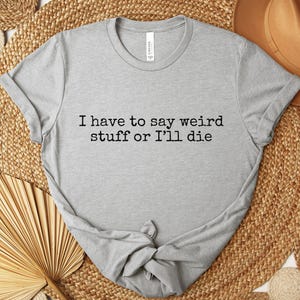 May include: A light gray t-shirt with the text "I have to say weird stuff or I'll die" in black. The shirt is knotted at the bottom and rests on a woven mat with a brown hat and decorative fan.