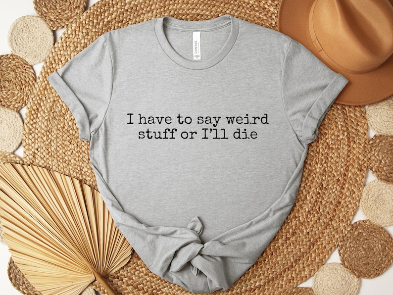 May include: A light gray t-shirt with the text "I have to say weird stuff or I'll die" in black. The shirt is knotted at the bottom and rests on a woven mat with a brown hat and decorative fan.