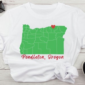 Puede incluir: Camiseta blanca con un contorno verde del estado de Oreg&oacute;n y un coraz&oacute;n rojo que marca la ubicaci&oacute;n de Pendleton, Oreg&oacute;n. El texto "Pendleton, Oreg&oacute;n" est&aacute; impreso debajo del mapa.