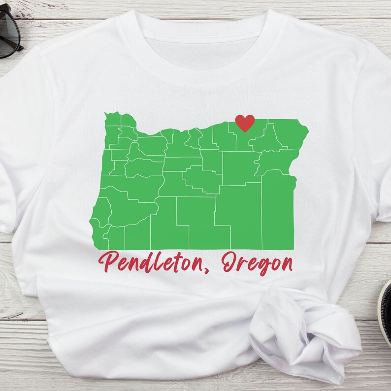 Puede incluir: Camiseta blanca con un contorno verde del estado de Oreg&oacute;n y un coraz&oacute;n rojo que marca la ubicaci&oacute;n de Pendleton, Oreg&oacute;n. El texto "Pendleton, Oreg&oacute;n" est&aacute; impreso debajo del mapa.