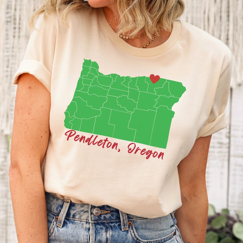 Puede incluir: Una camiseta beige con un contorno verde del estado de Oreg&oacute;n y un coraz&oacute;n rojo que marca la ubicaci&oacute;n de Pendleton, Oreg&oacute;n. El texto "Pendleton, Oreg&oacute;n" est&aacute; escrito en rojo debajo del mapa.