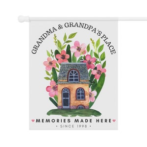 May include: A white garden flag with a house surrounded by pink flowers and green leaves. The text reads "Grandma & Grandpa's Place" in a circle around the house and "Memories Made Here. Since 1998." at the bottom.