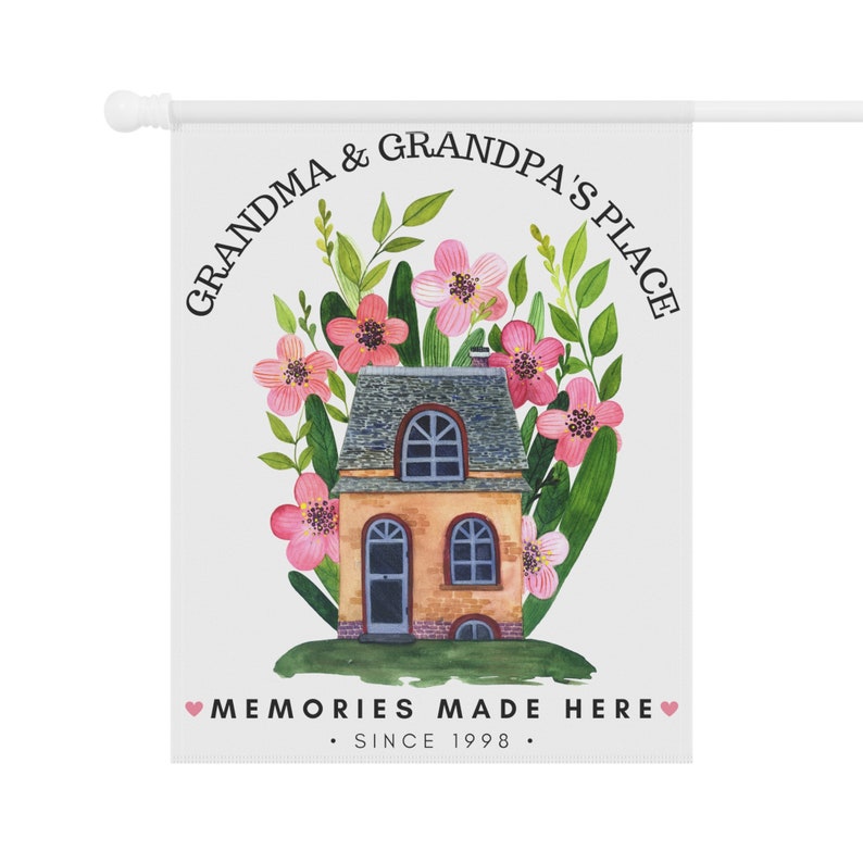 May include: A white garden flag with a house surrounded by pink flowers and green leaves. The text reads "Grandma & Grandpa's Place" in a circle around the house and "Memories Made Here. Since 1998." at the bottom.