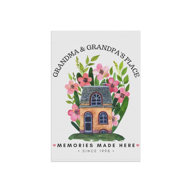 May include: A white garden flag with a watercolor illustration of a house surrounded by pink flowers and green leaves. The text "Grandma & Grandpa's Place" is in a circle above the house and the text "Memories Made Here. Since 1998." is below the house.