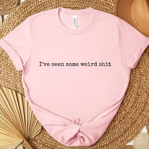 May include: A light pink t-shirt with the phrase "I've seen some weird shit" printed in black. The shirt is knotted at the bottom and rests on a woven mat with a brown hat and dried palm leaves.