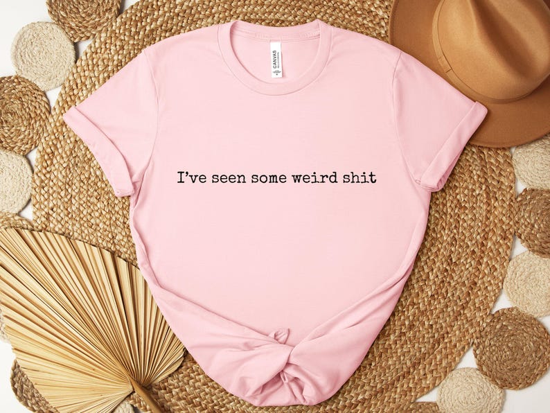 May include: A light pink t-shirt with the phrase "I've seen some weird shit" printed in black. The shirt is knotted at the bottom and rests on a woven mat with a brown hat and dried palm leaves.