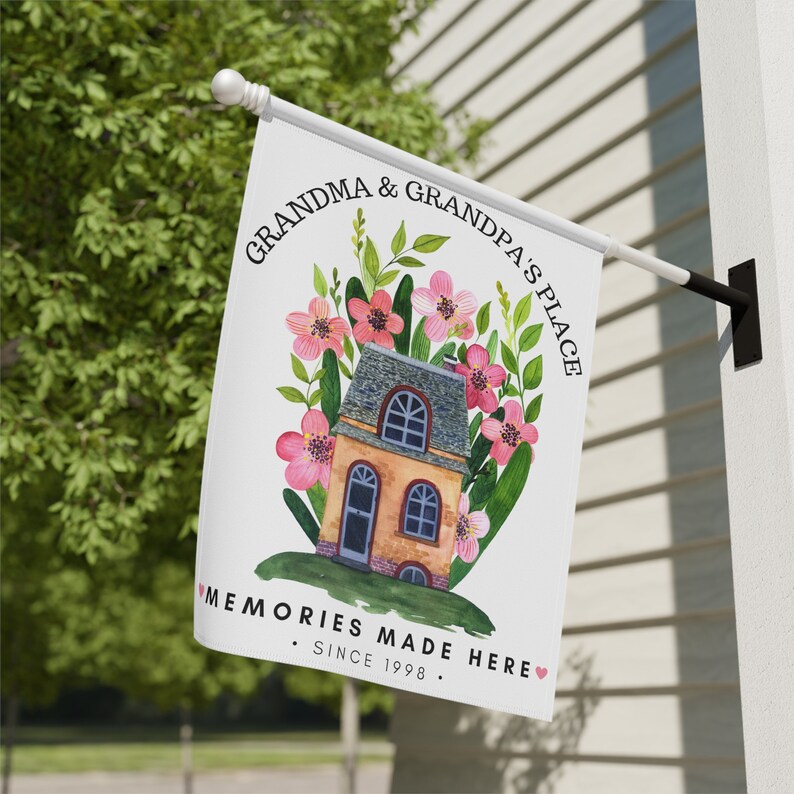 May include: A white garden flag with a house illustration surrounded by pink flowers and green leaves. The text reads "Grandma & Grandpa's Place Memories Made Here. Since 1998."