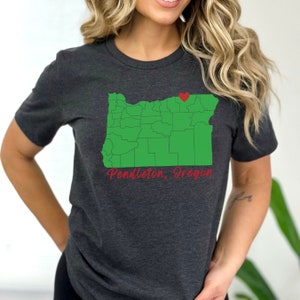Puede incluir: Una camiseta gris oscuro con un contorno verde del estado de Oreg&oacute;n y un coraz&oacute;n rojo que marca la ubicaci&oacute;n de Pendleton, Oreg&oacute;n. El texto "Pendleton, Oreg&oacute;n" est&aacute; escrito en rojo cursivo debajo del mapa.