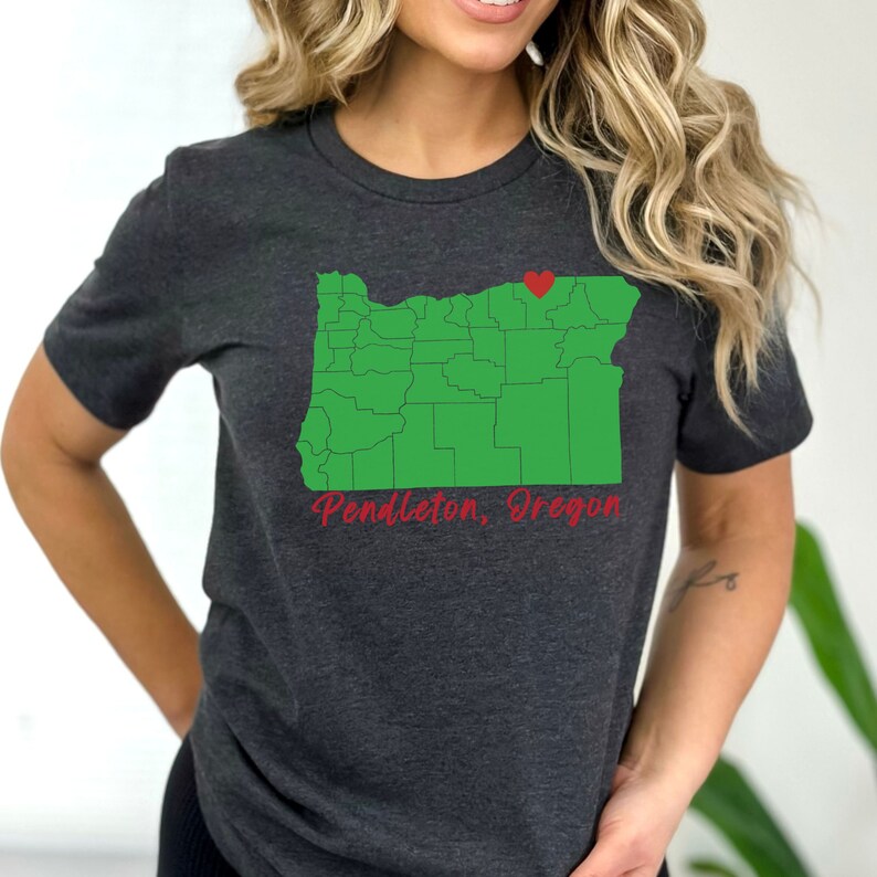 Puede incluir: Una camiseta gris oscuro con un contorno verde del estado de Oreg&oacute;n y un coraz&oacute;n rojo que marca la ubicaci&oacute;n de Pendleton, Oreg&oacute;n. El texto "Pendleton, Oreg&oacute;n" est&aacute; escrito en rojo cursivo debajo del mapa.
