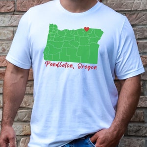 Puede incluir: Camiseta blanca con un contorno verde del estado de Oreg&oacute;n y un coraz&oacute;n rojo sobre Pendleton, Oreg&oacute;n. El texto "Pendleton, Oreg&oacute;n" est&aacute; escrito en rojo debajo del contorno del estado.