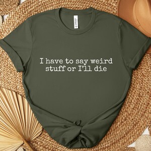 May include: Olive green t-shirt with the text "I have to say weird stuff or I'll die" in white. The shirt is knotted at the bottom and is displayed on a woven surface with a brown hat.