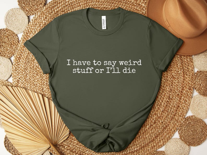 May include: Olive green t-shirt with the text "I have to say weird stuff or I'll die" in white. The shirt is knotted at the bottom and is displayed on a woven surface with a brown hat.