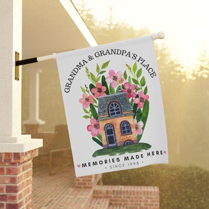 May include: A white flag with a watercolor illustration of a house with pink flowers and green leaves. The text on the flag reads "Grandma & Grandpa's Place. Memories Made Here. Since 1998."