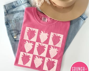 May include: Pink t-shirt with a white graphic of strawberries in a checkered pattern. The shirt is laid out on a blue denim background.