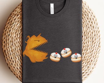 May include: Dark gray t-shirt featuring a cartoon design. The design shows a Pac-Man-like character chasing three sufganiyot, a type of jelly donut. The shirt is made of a soft material and has a crew neck.