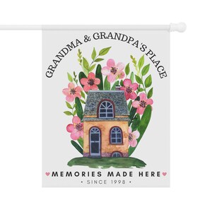 May include: A white garden flag with a watercolor illustration of a house surrounded by pink flowers and green leaves. The text reads "Grandma & Grandpa's Place" in a circle around the house and "Memories Made Here. Since 1998." at the bottom.