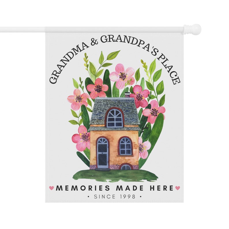 May include: A white garden flag with a watercolor illustration of a house surrounded by pink flowers and green leaves. The text reads "Grandma & Grandpa's Place" in a circle around the house and "Memories Made Here. Since 1998." at the bottom.