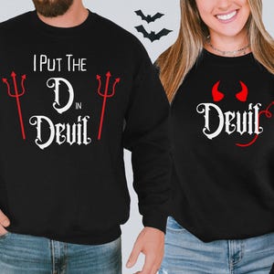 May include: Two black sweatshirts with a Halloween theme. One sweatshirt says "I put the D in Devil" with red devil horns and a red trident. The other sweatshirt says "Devil" with red devil horns.