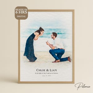 Puede incluir: Impresión acuarela enmarcada que representa una propuesta de matrimonio en la playa. Un hombre arrodillado ofrece un anillo a una mujer con un vestido azul largo. Texto: "CHLOE & LIAM" y "In the light of day, I said yes to forever. June 14, 2025."