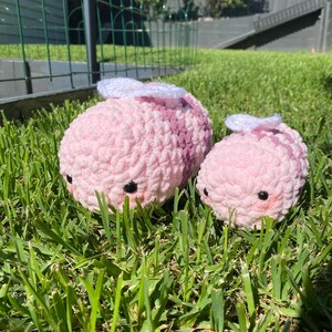 May include: Two pink and purple crocheted bee plushies with white wings. The larger bee is in the foreground and the smaller bee is behind it. Both bees have black eyes and are sitting on green grass.