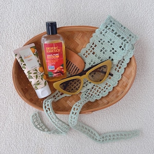 Puede incluir: Una colecci&oacute;n de productos de belleza y cuidado personal dispuestos en una cesta de mimbre. Incluye un tubo de crema de manos, una botella de aceite de jojoba, un peine de madera, gafas de sol amarillas de ojo de gato y una diadema de ganchillo verde claro.