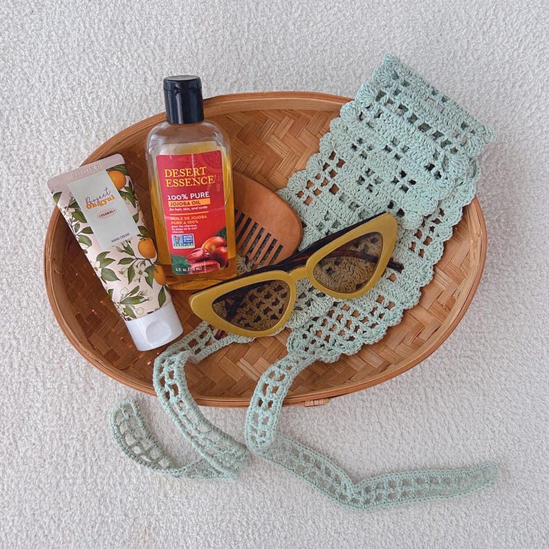 Puede incluir: Una colecci&oacute;n de productos de belleza y cuidado personal dispuestos en una cesta de mimbre. Incluye un tubo de crema de manos, una botella de aceite de jojoba, un peine de madera, gafas de sol amarillas de ojo de gato y una diadema de ganchillo verde claro.