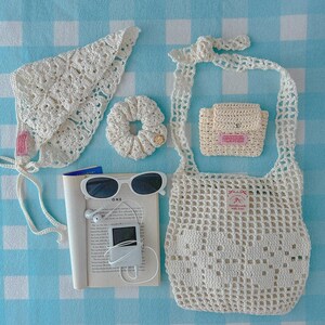 May include: A collection of cream-colored crochet accessories including a triangular headscarf, a hair scrunchie, a small pouch, and a net tote bag. Also included are white sunglasses, a white digital music player with earphones, and an open book.