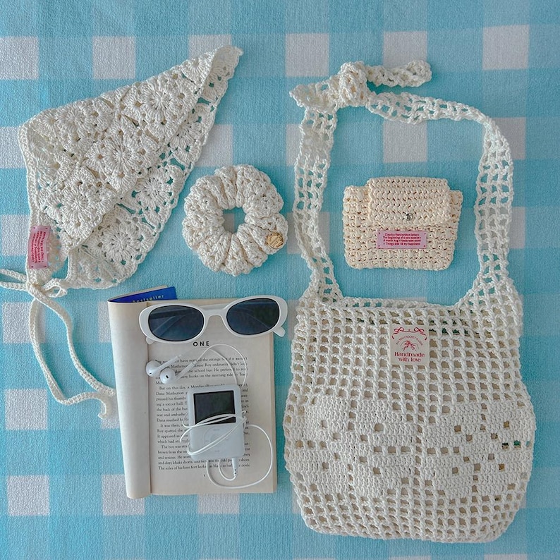 May include: A collection of cream-colored crochet accessories including a triangular headscarf, a hair scrunchie, a small pouch, and a net tote bag. Also included are white sunglasses, a white digital music player with earphones, and an open book.