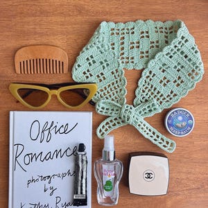 Puede incluir: Una variedad de art&iacute;culos sobre una superficie de madera. Incluye un peine de madera, gafas de sol amarillas de ojo de gato, un libro titulado "Office Romance", un tubo de pintura a rayas blancas y negras, una bufanda de cuello de ganchillo verde, una botella de fragancia "Body Fantasies" French Pear, una peque&ntilde;a lata redonda de "Badger Sleep Balm" y un estuche.