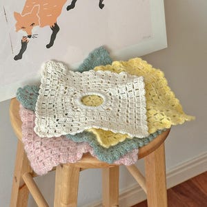 Pode incluir: Um conjunto de bases de copos de crochê feitas à mão em cores pastel: branco, rosa, amarelo e verde-azulado. Cada base de copo tem uma borda ondulada e uma abertura oval no centro. Empilhadas em um banquinho de madeira.