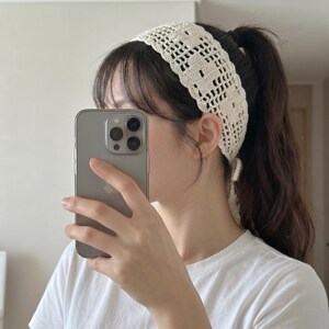 Puede incluir: Diadema de ganchillo color crema con un patr&oacute;n geom&eacute;trico. La diadema es ancha y se envuelve alrededor de la cabeza. La imagen muestra a una persona que lleva la diadema con una camiseta blanca y sosteniendo un tel&eacute;fono inteligente.