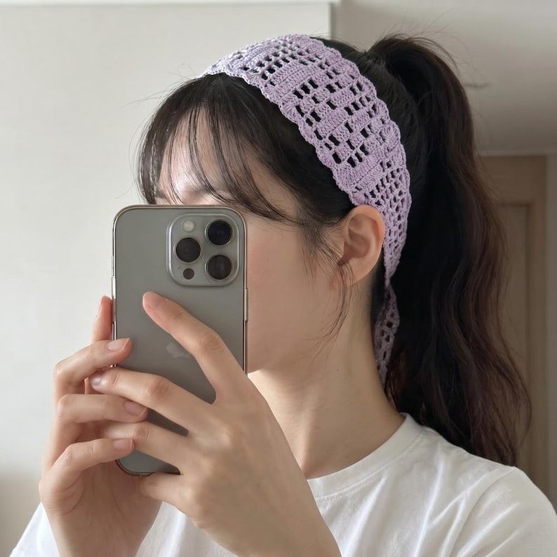 Puede incluir: Una diadema de ganchillo de color lila claro con un dise&ntilde;o calado rectangular. La diadema se lleva en la cabeza y se ata en la parte posterior. La persona de la imagen sostiene un smartphone plateado.